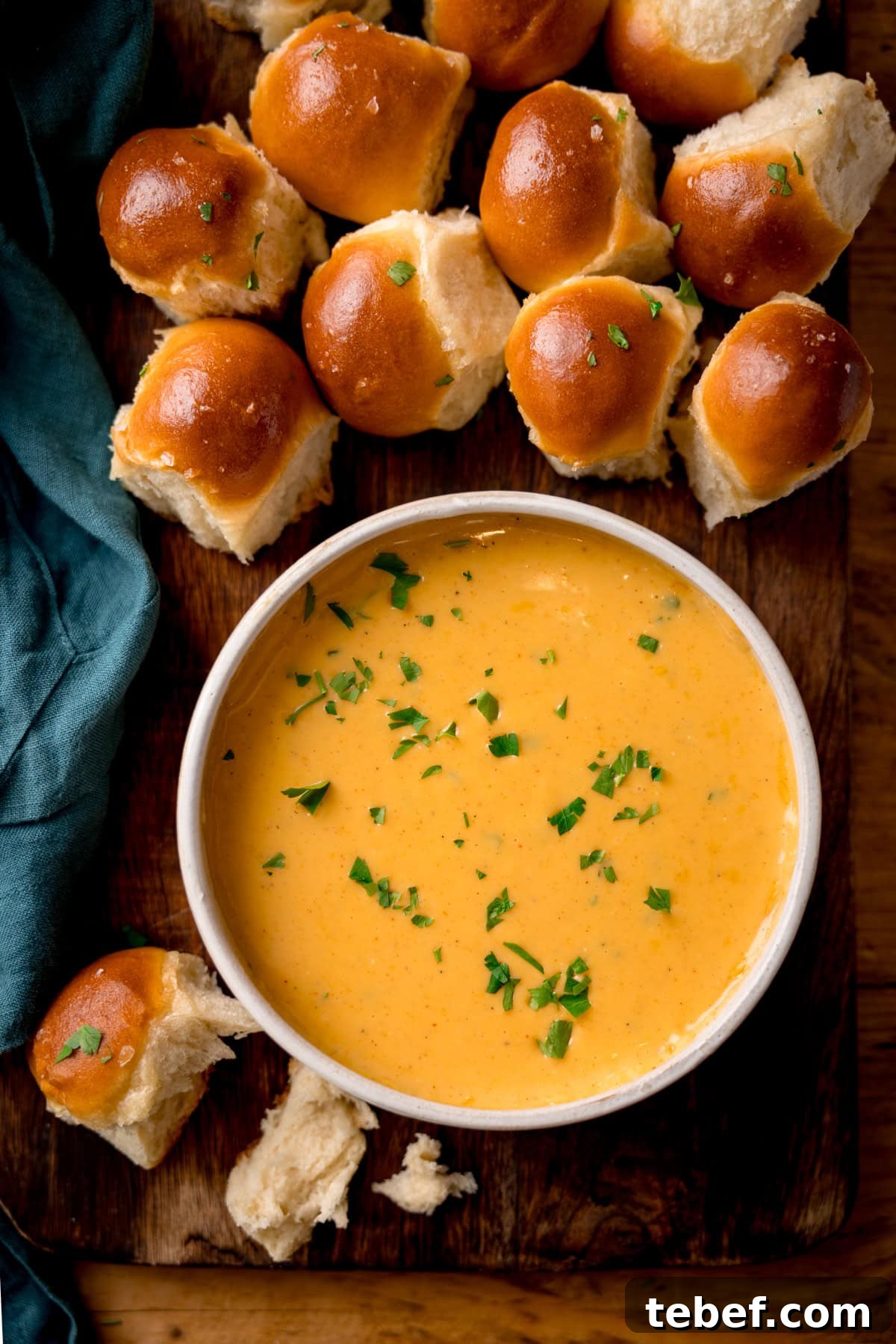 Creamy Beer Cheese Dip Recipe for Game Day and Parties 4 A bowl of beer cheese dip is garnished with chopped herbs on a wooden board. Surrounding it are several soft, shiny dough balls. A teal cloth is on from the left-hand side of the board.