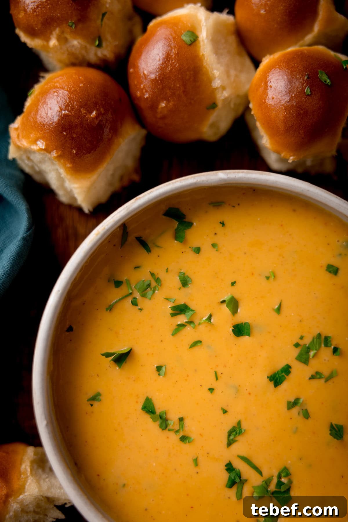 Creamy Beer Cheese Dip Recipe for Game Day and Parties 5 A bowl beer cheese dip is garnished with chopped herbs and sits on a wooden table. Surrounding the bowl are several shiny dough balls. A corner of a teal napkin is visible to the left.
