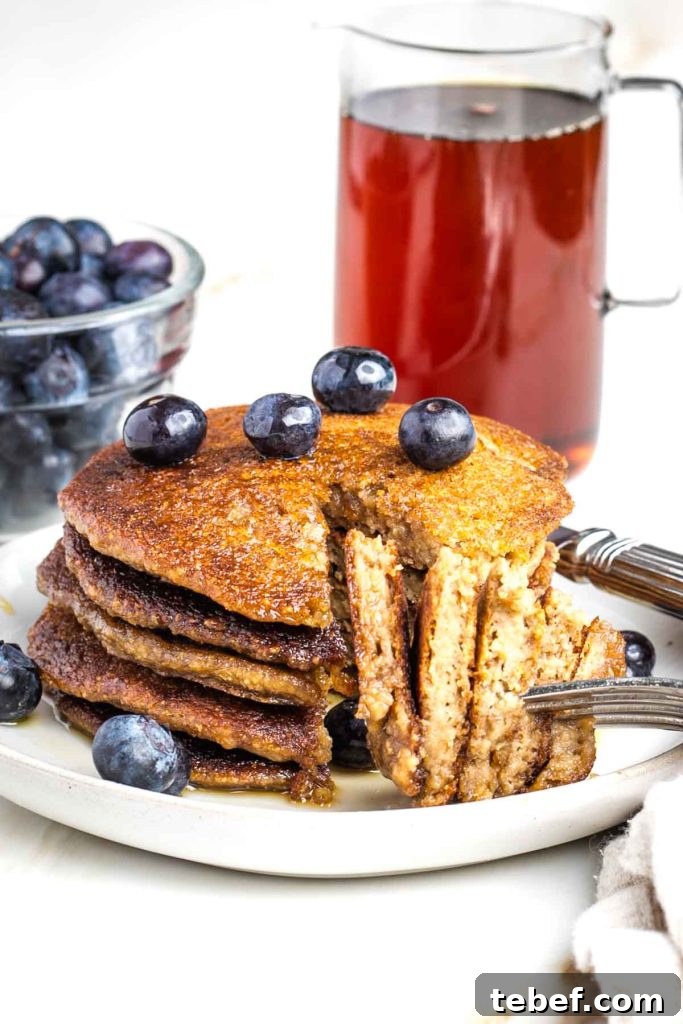 Stack of banana oat pancakes with a bite cut and ready to go on a fork. Blueberries and maple syrup in the background.