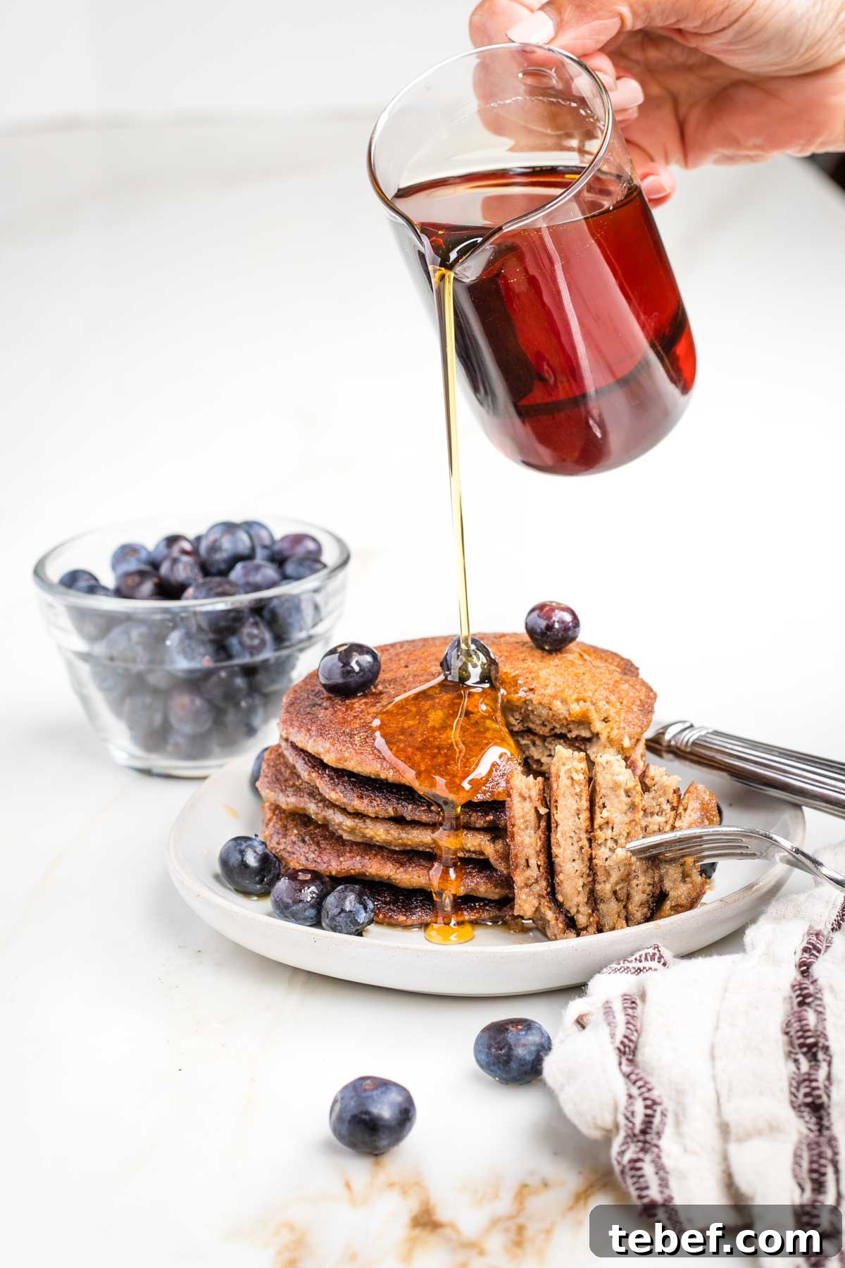 A bite cut out of a stack of banana oatmeal pancakes with maple syrup being poured over the plate.