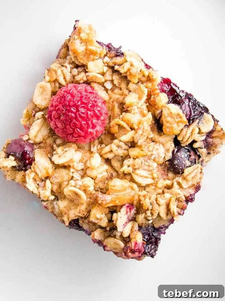 Close up of a square of baked oatmeal, topped with a bright red raspberry and some blueberries plus oats and nuts.