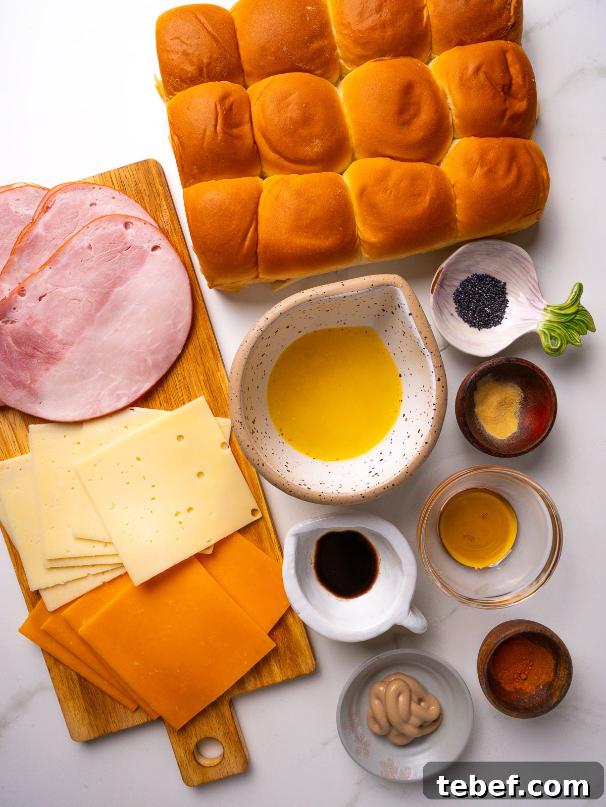 Overhead view of ingredients for Hawaiian ham and cheese sliders: sweet Hawaiian rolls, deli ham, Swiss and cheddar, butter, Dijon, Worcestershire and poppy seeds.
