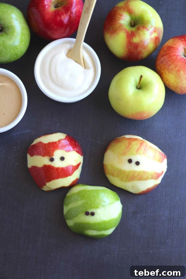 3 apple mummies on black surface with whole apples and small dishes of fruit dips nearby.
