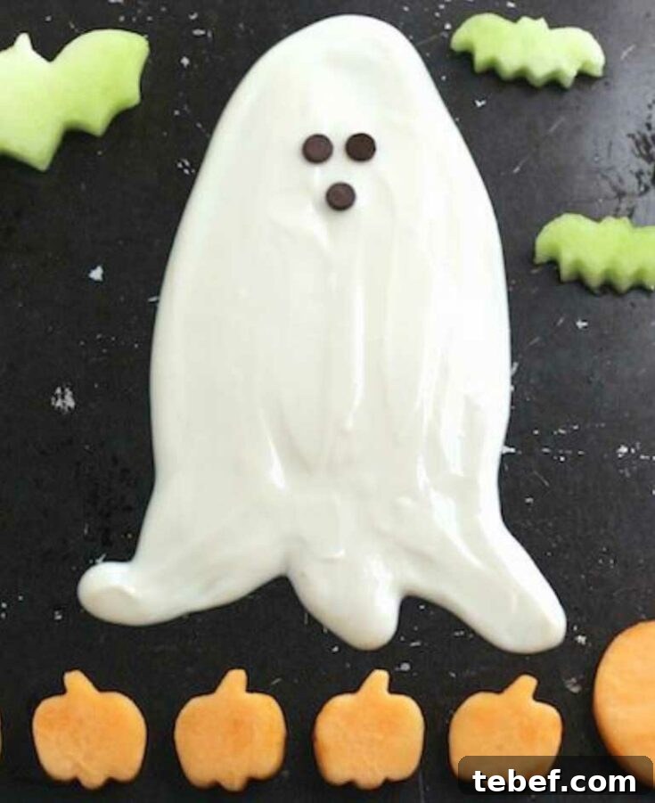 Fruit dip shaped like ghost on black surface surrounded by fruit cut in shapers of pumpkins and bats.