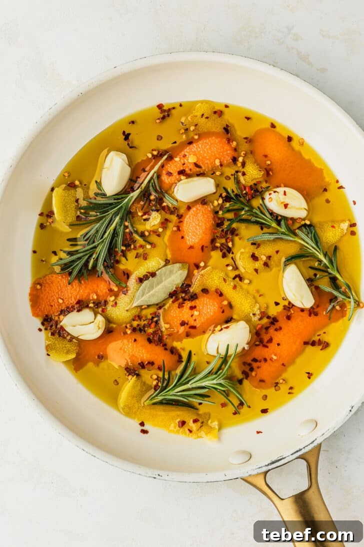 Citrus-Marinated Spanish Olives with Manchego Cheese 4 A white pan of olive oil, orange peels, lemon peels, garlic, and rosemary sprigs on a tan counter.