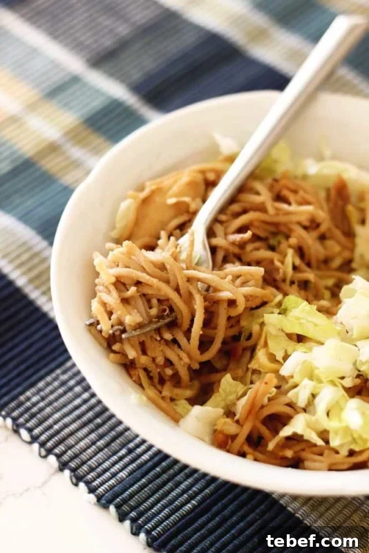 White bowl containing White bowl containing Thai Peanut Chicken Pasta, sitting on a blue placement, silver fork in noodles.