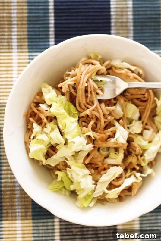 White bowl containing Thai Peanut Chicken Pasta, sitting on a blue placement map topped with cabbage, silver fork in noodles.