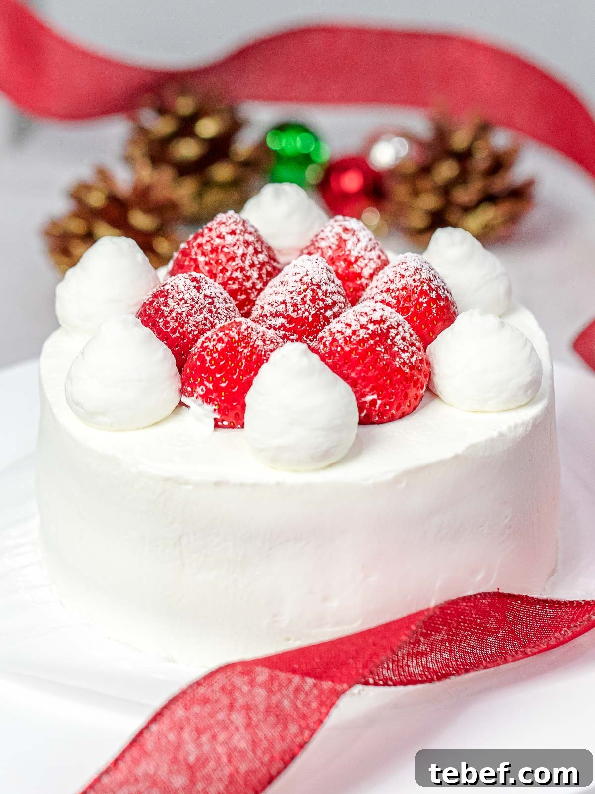 Authentic Japanese Strawberry Shortcake Recipe — Light, Fluffy Layers 4 Japanese Christmas cake decorated with whole strawberries dusted with powdered sugar with Christmas ornaments, red ribbon, and pine cones in the back