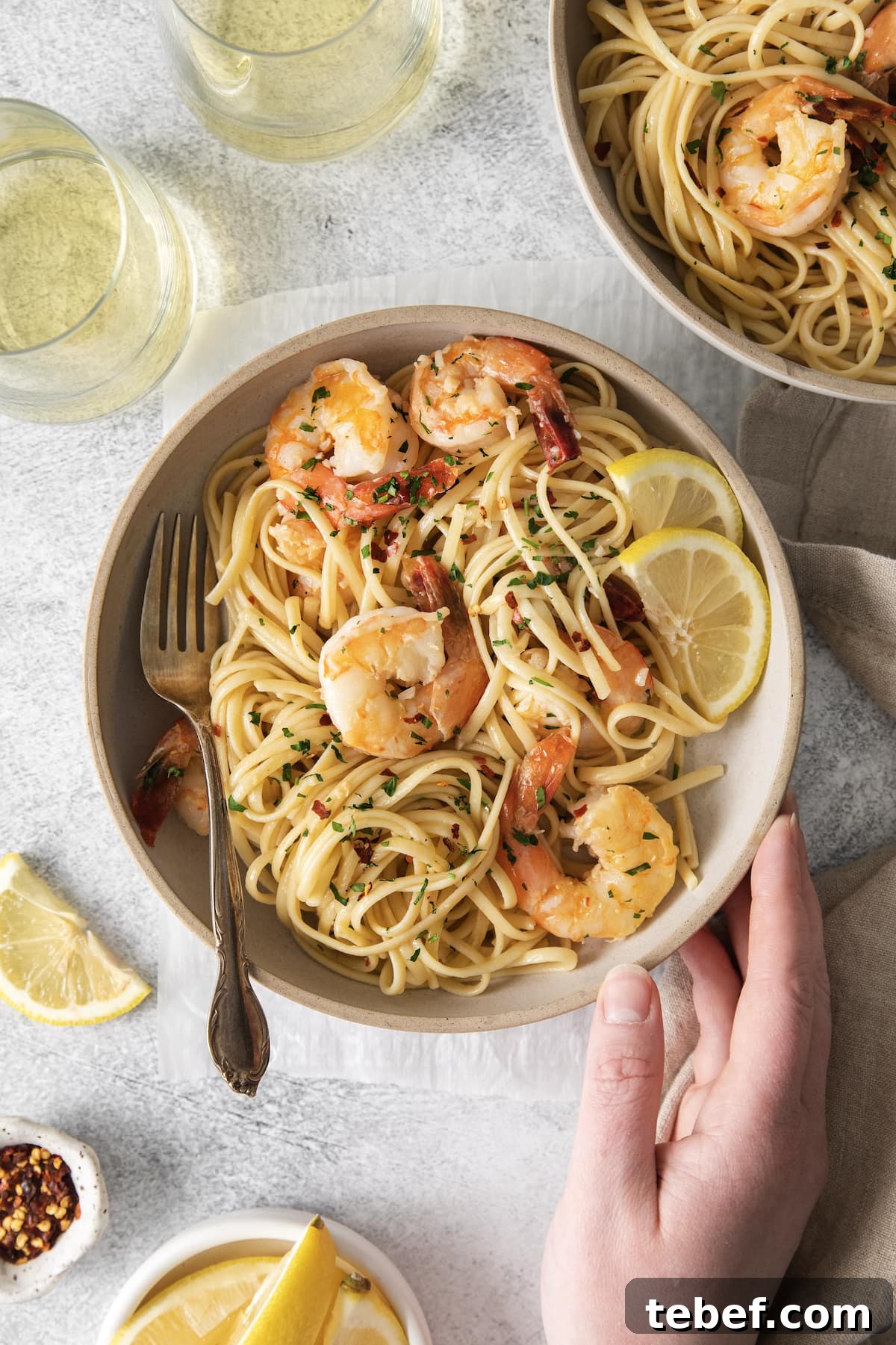 Garlic Butter Shrimp Scampi Recipe for Quick Weeknight Meals 3 a hand touching a bowl with shrimp pasta with linguine, red pepper flakes, herbs, and lemon slices