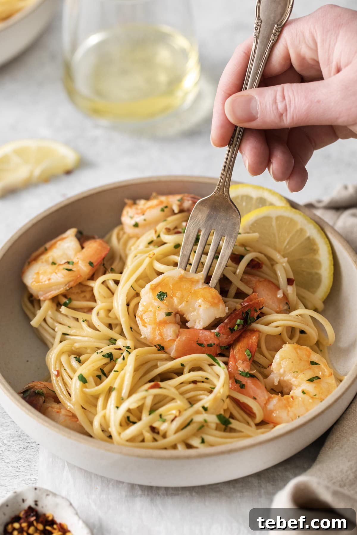 Garlic Butter Shrimp Scampi Recipe for Quick Weeknight Meals 10 a hand holding a fork twirling shrimp pasta with linguine, red pepper flakes, herbs, and lemon slices in a bowl