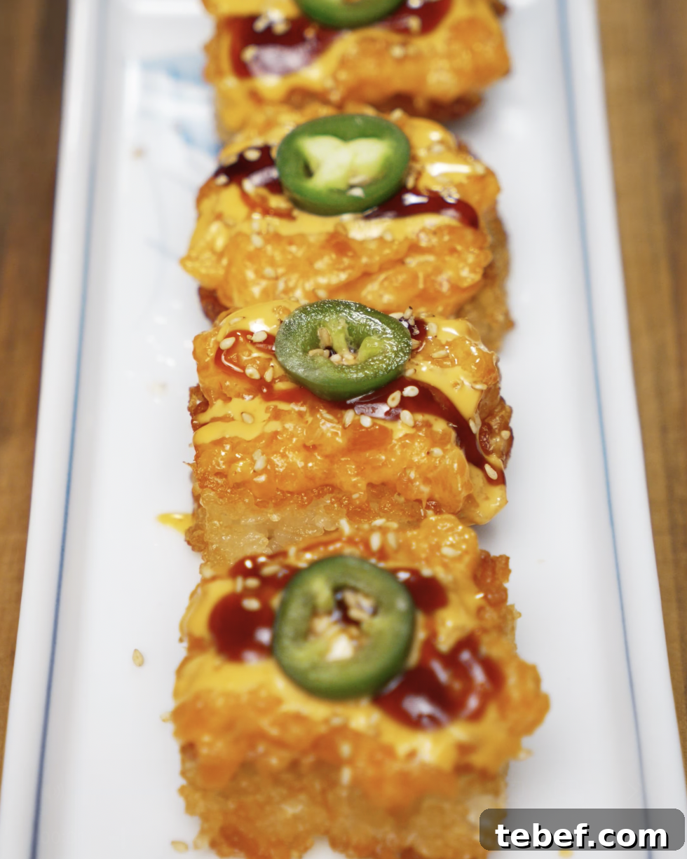 Rows of crispy rice squares topped with spicy salmon mix, spicy mayo, sweet soy glaze, and fresh jalapeño slices. Finished with sesame seeds and plated on a rectangular white dish.