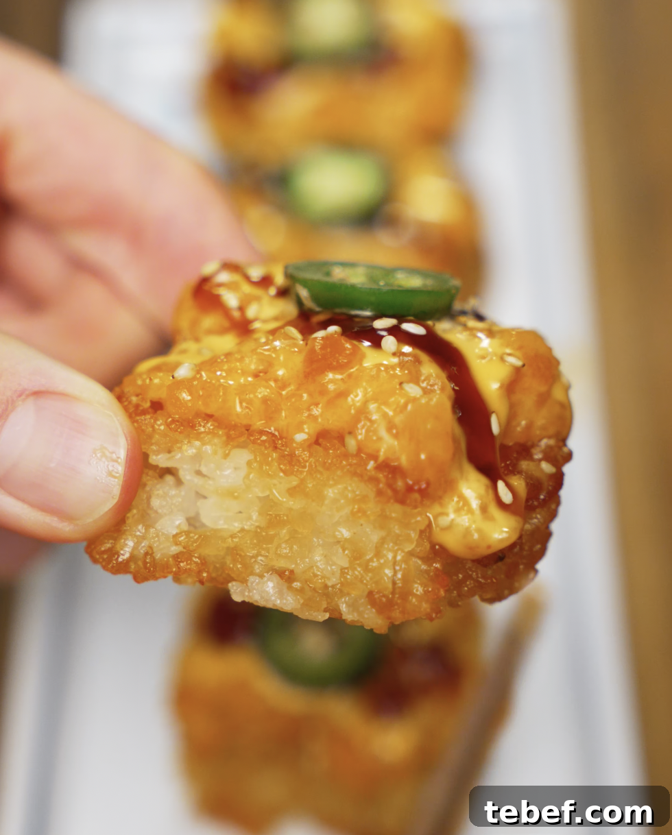 A hand holding a crispy rice bite topped with creamy, spicy salmon mixture, jalapeño, and sesame seeds. The texture of the crispy rice and the juicy salmon topping is clearly visible.