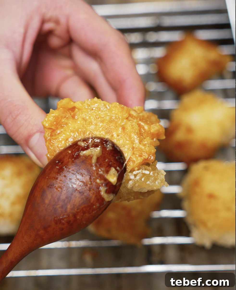 A hand holding a crispy rice cube while brushing spicy salmon on top with a wooden spoon. The rice is perfectly fried, showcasing its crunchy texture before assembling the salmon bites.