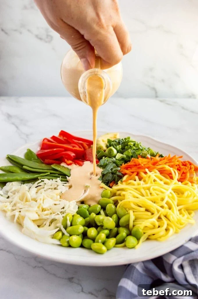 A hand pouring creamy light brown liquid from a small glass jug over a plate of vegetables.
