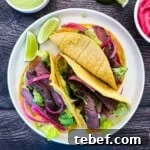 Two seared Wagyu steak tacos in corn tortillas topped with pickled red onions, shredded lettuce, and avocado lime crema, served on a white plate with lime wedges.