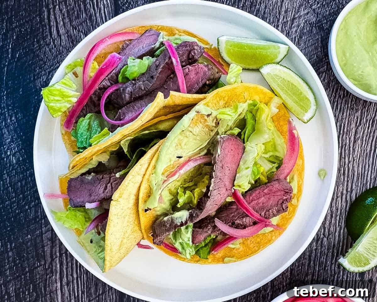 Three steak tacos on a white dinner plate, topped with avocado lime crema and pickled red onions.