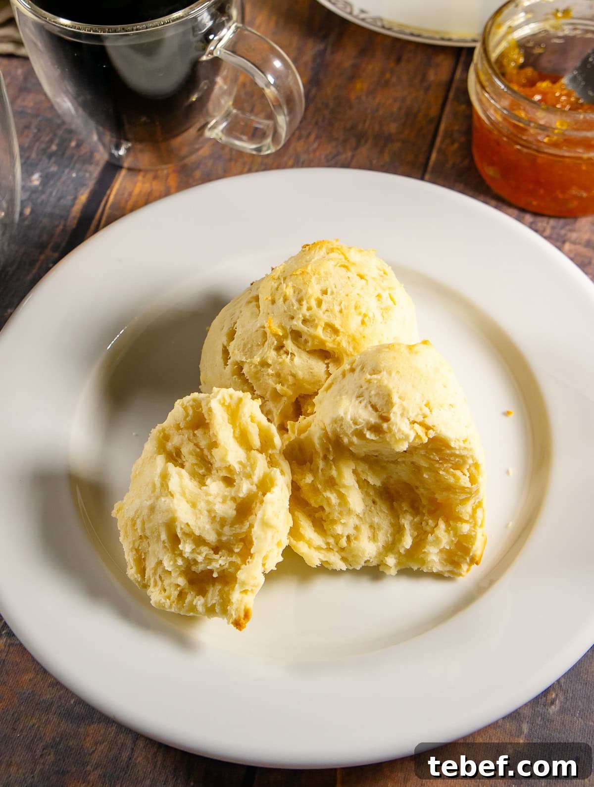Flaky No-Knead Drop Biscuits Recipe for Quick Homemade Baking 2 fluffy no-knead biscuit cut open on a white plate with a jar of orange jam and a cup of coffee beside it.