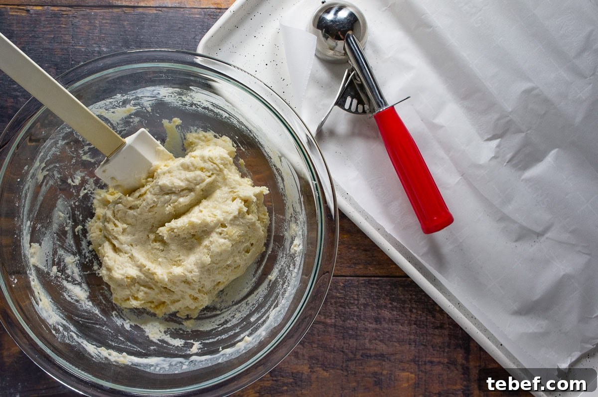 Flaky No-Knead Drop Biscuits Recipe for Quick Homemade Baking 4 no-knead biscuit dough in a bowl with a cookie scoop on a baking sheet beside it.