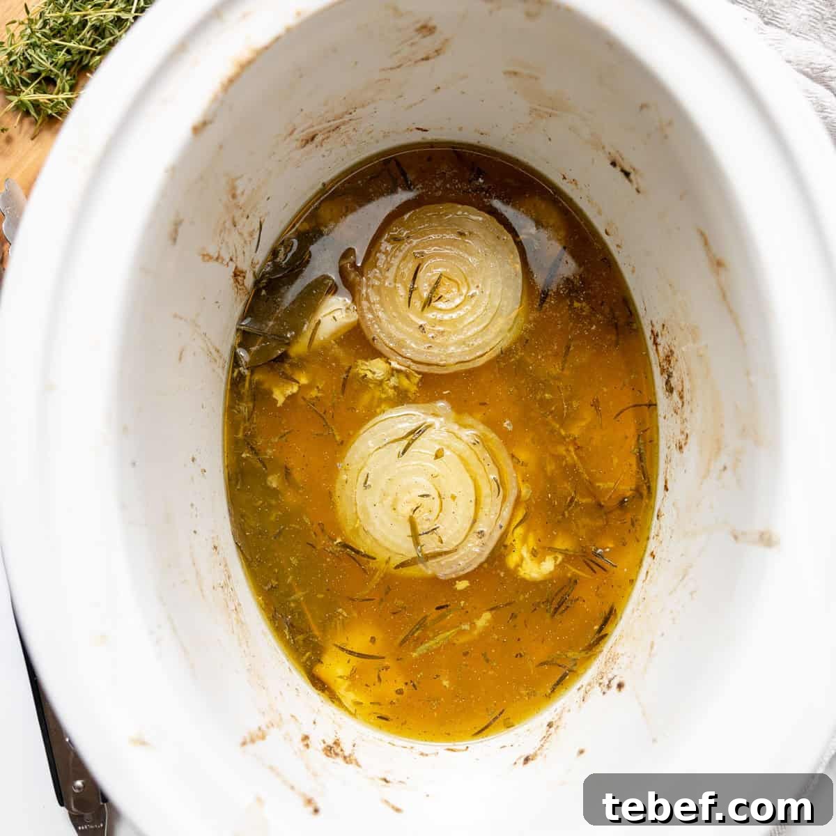 A slow cooker filled with broth, herbs, and two halved onions.