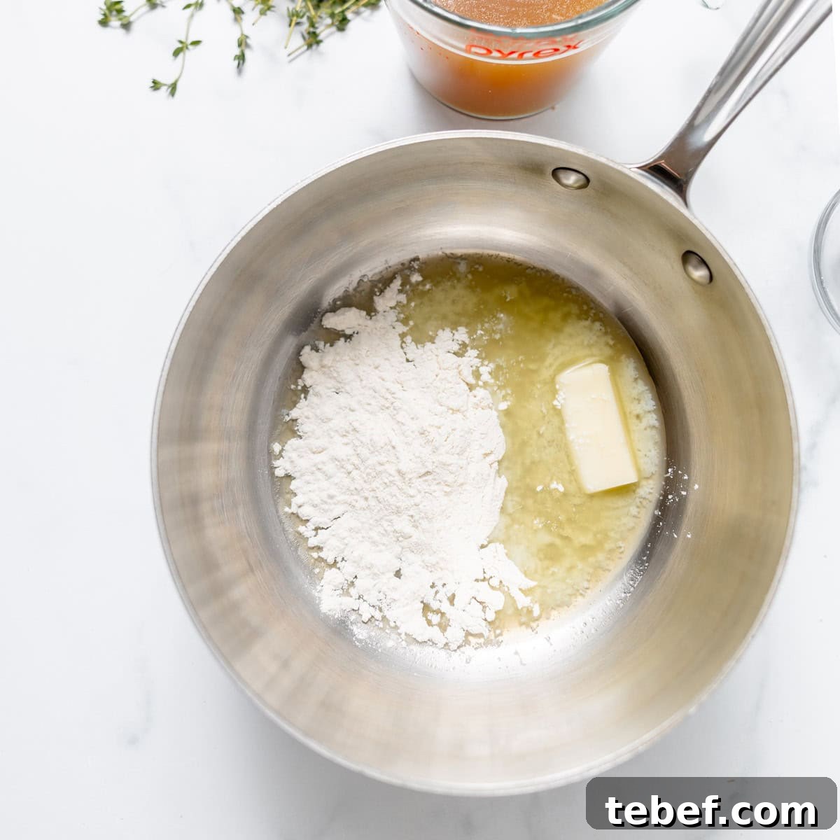 A saucepan with melted butter and flour, ready to be mixed, sits on a white countertop beside a glass measuring cup and some sprigs of thyme.