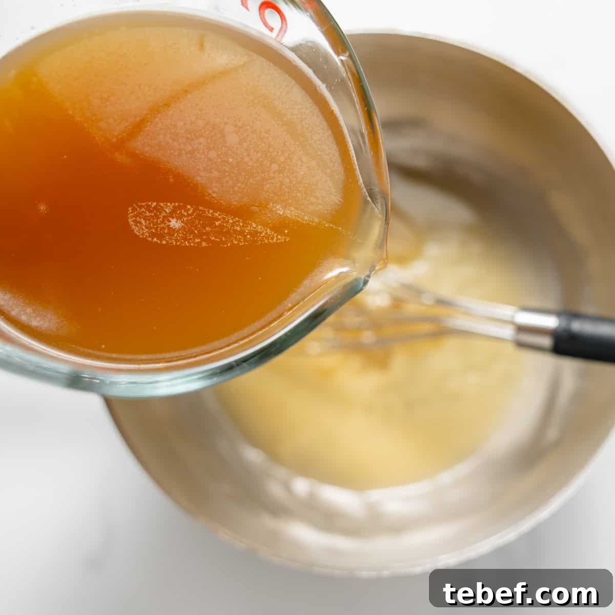 A glass measuring cup of brown liquid is being poured into a metal bowl with a whisk and a light-colored mixture inside.