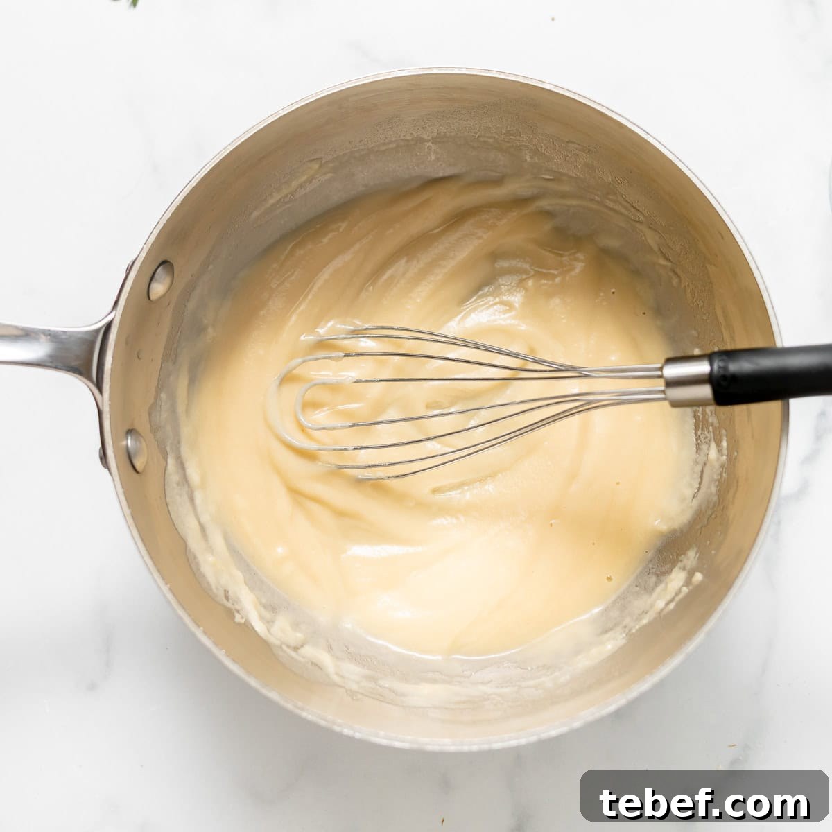 A whisk resting in a saucepan containing a smooth, thick, pale mixture on a white surface.