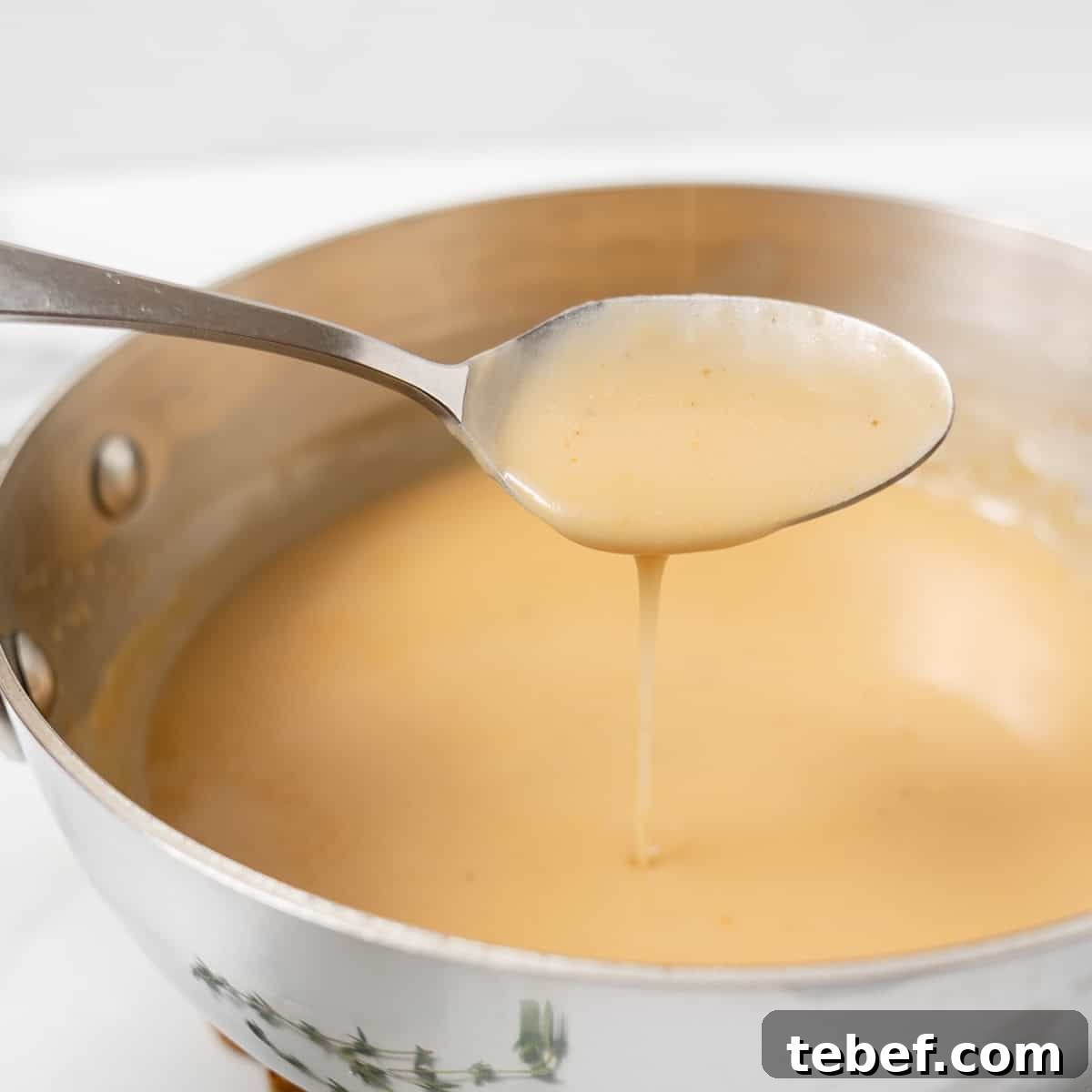 A spoon holds up a stream of light brown gravy above a stainless steel pan filled with more of the sauce.