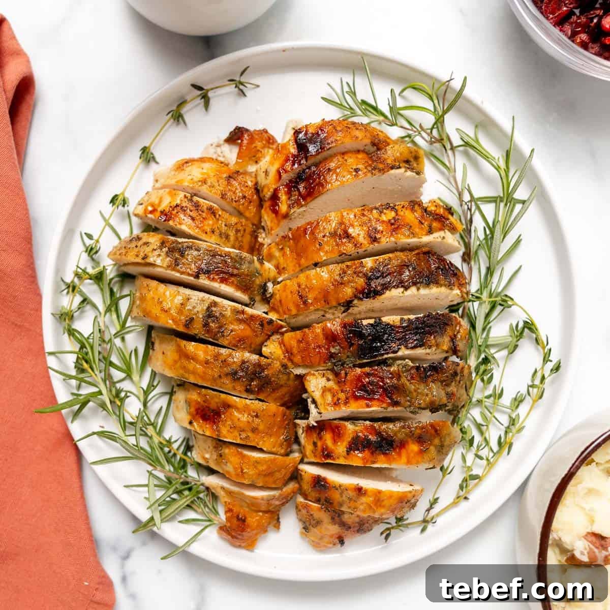 Sliced roasted turkey breast arranged on a white plate with sprigs of rosemary and thyme, set on a white marble surface.