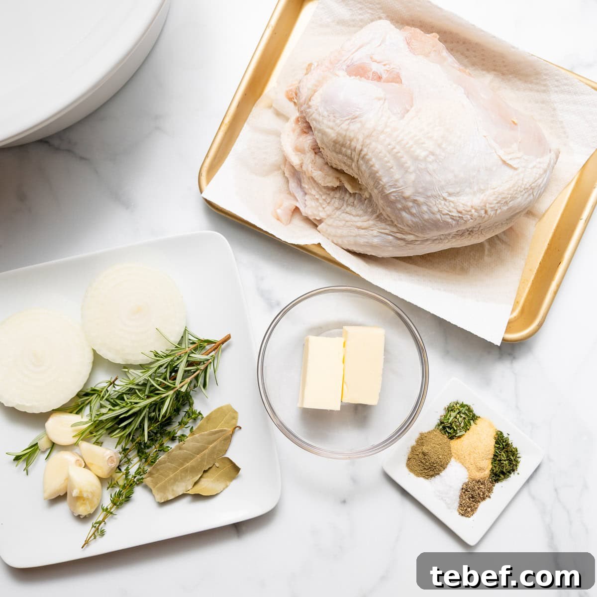 Raw turkey breast on a tray, sliced onions, garlic, fresh herbs, and bay leaves on a plate, butter in a bowl, and assorted dried spices on a small dish, arranged on a white surface.
