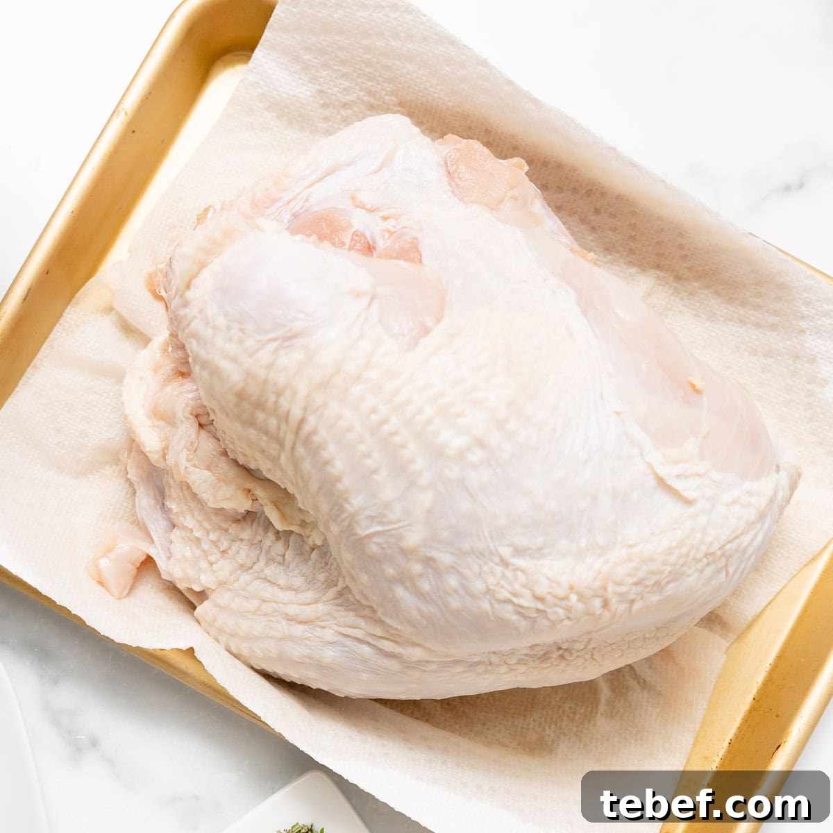 Raw turkey breast placed on paper towels in a gold baking tray, ready for preparation, on a white marble surface.
