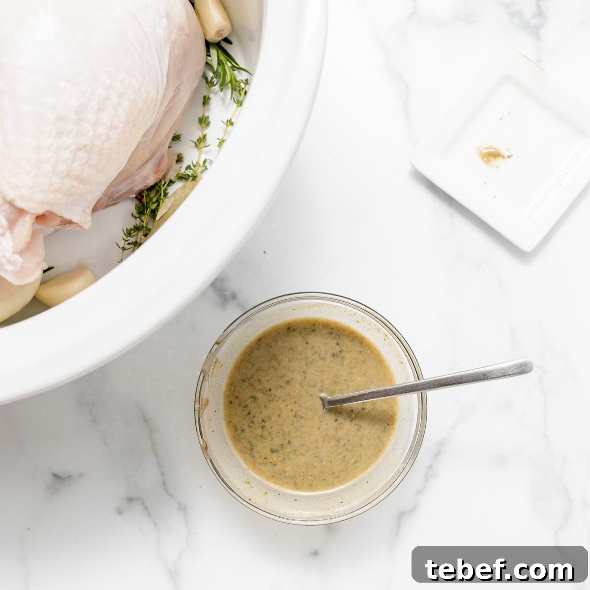 A bowl of seasoned butter with a spoon next to a raw turkey breast, herbs, and garlic in a white dish on a marble countertop.