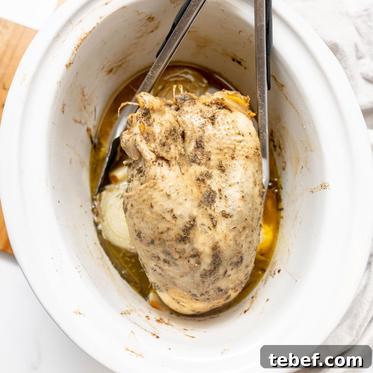 Cooked seasoned turkey breast being lifted from a white slow cooker with metal tongs, surrounded by broth and onion slices.