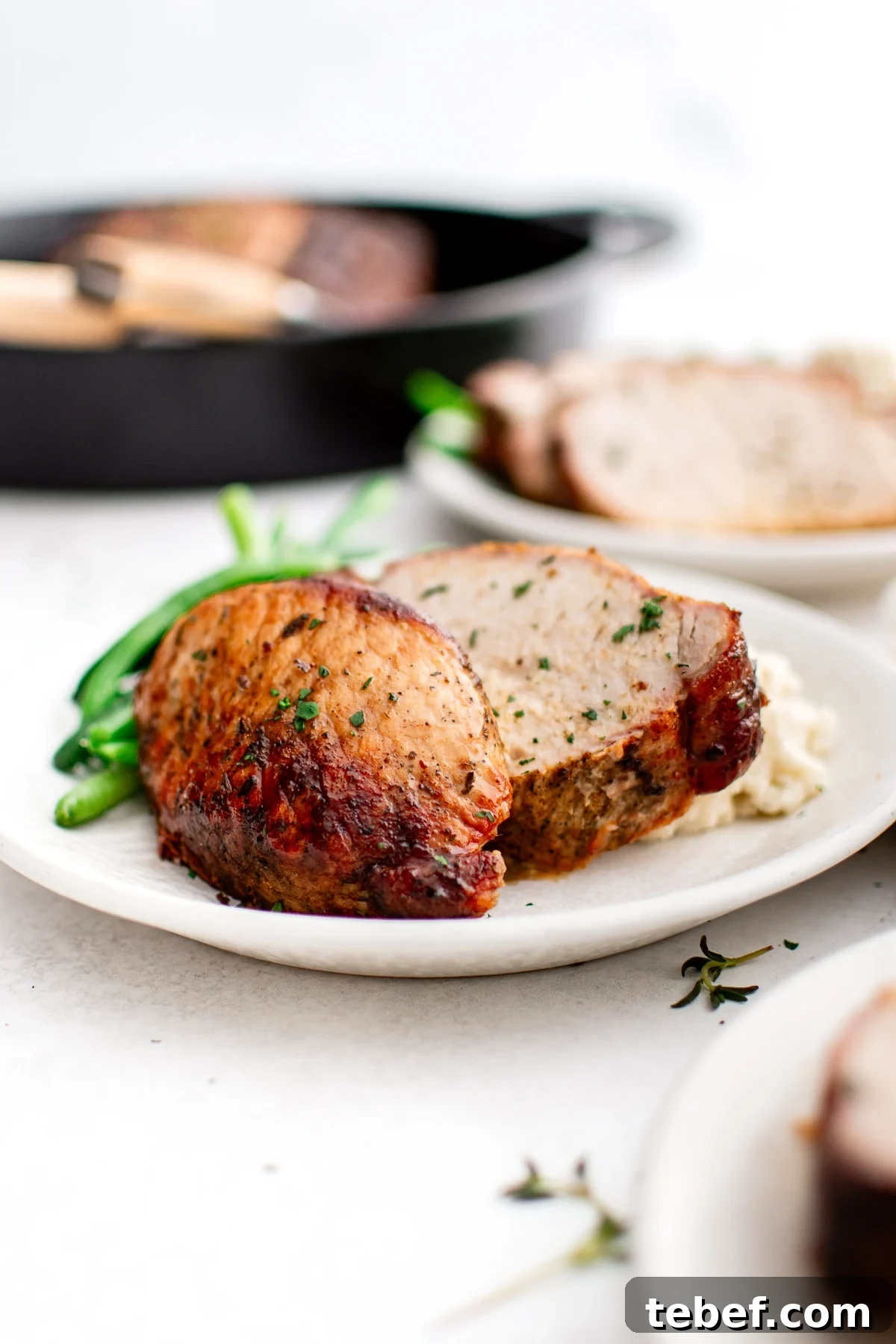 Perfect Oven-Roasted Pork Loin: Juicy, Crispy Recipe and Tips 7 Two slices of juicy pork loin are plated with creamy mashed potatoes and fresh green beans on a white dinner plate. In the background, the cast iron skillet with the remaining roast adds a rustic, home-cooked feel to the scene.