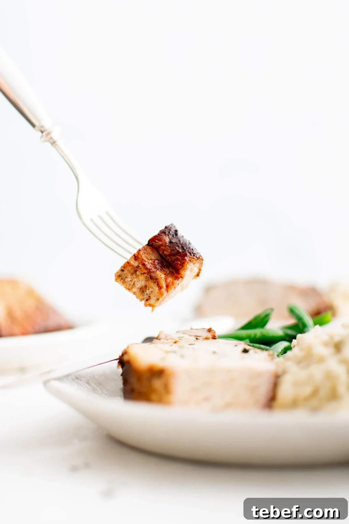 Perfect Oven-Roasted Pork Loin: Juicy, Crispy Recipe and Tips 8 A fork gently lifts a slice of juicy pork loin from a white plate, showcasing the tender texture and golden crust. Mashed potatoes and green beans are partially visible beneath, completing the comforting meal presentation.