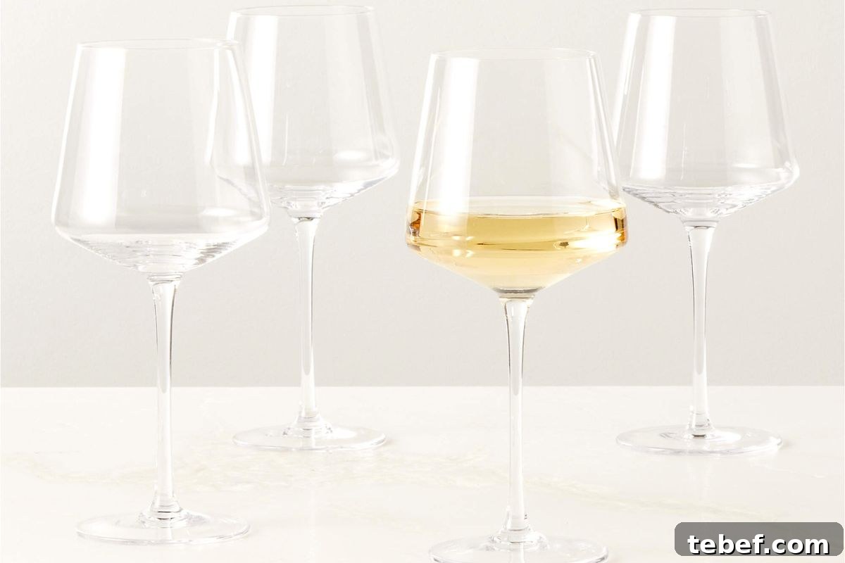 Four empty wine glasses on a white surface, with one glass partially filled with white wine.