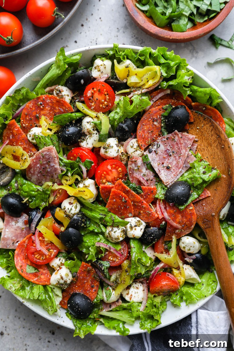 A serving bowl filled with loaded Italian salad surrounded by cherry tomatoes and a bowl of sliced fresh basil.